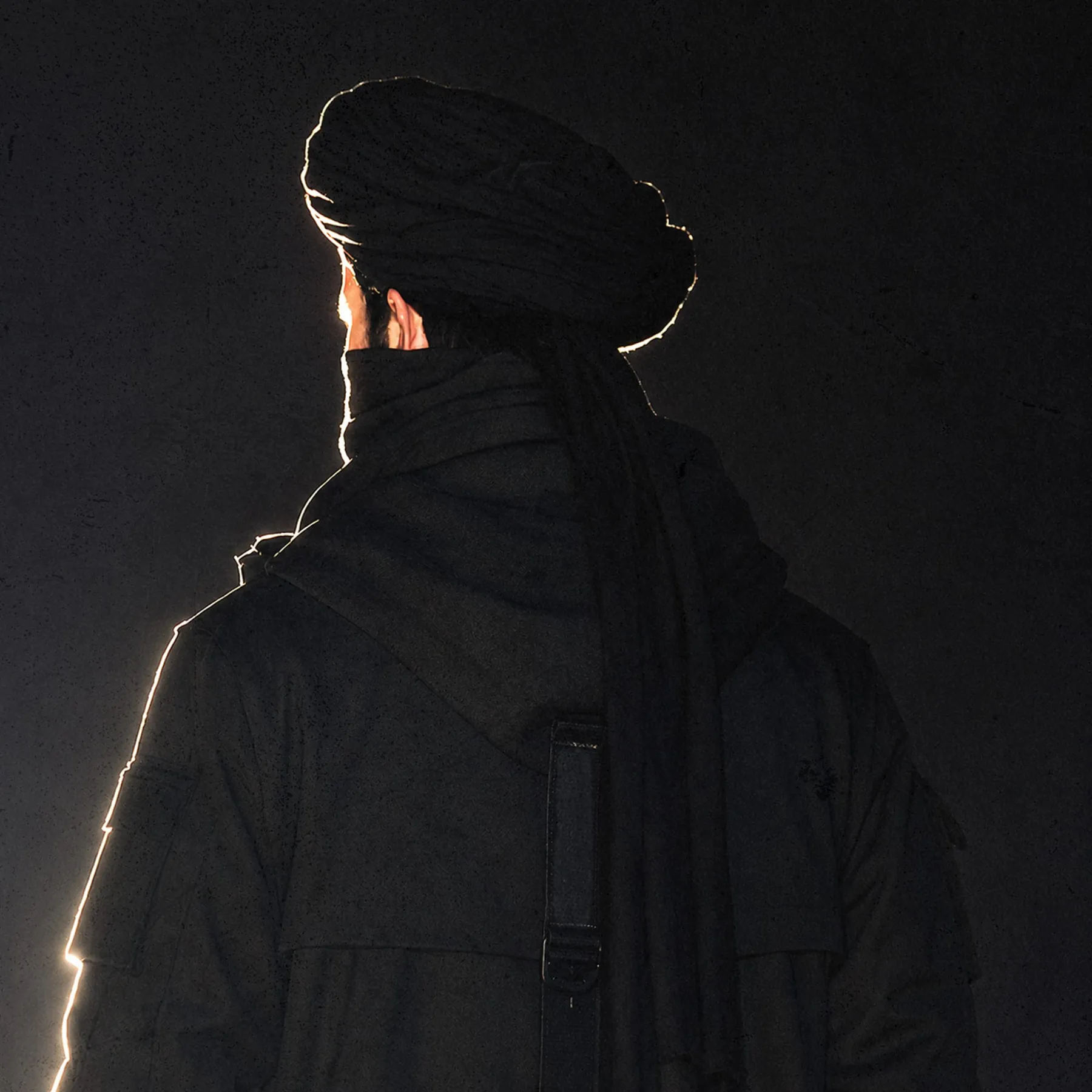 REBEL X from behind, in black, rim-lit against darkness — second study.