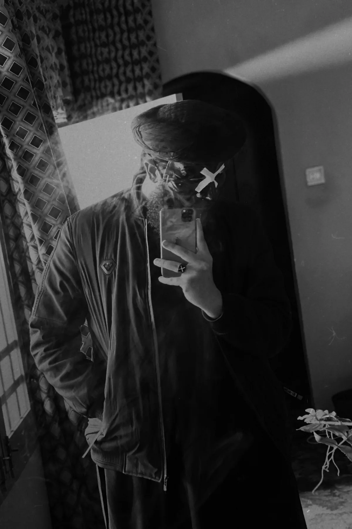 Mirror self-portrait: the poet in cap and mask marked with a white X, face hidden, phone in hand.